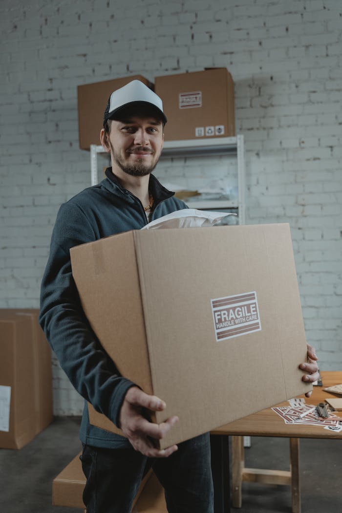 Smiling courier in cap carrying a fragile package in a warehouse setting.